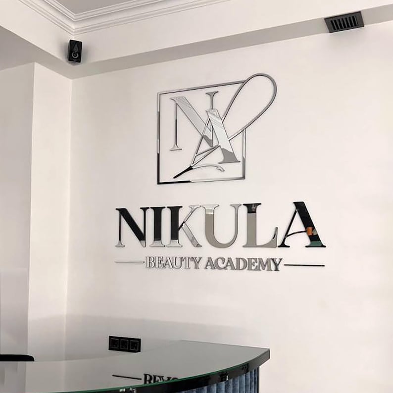 Może przedstawiać: Biała ściana z logo "Nikula Beauty Academy". Logo zawiera stylizowane "N" i "A" w kwadratowej ramie, z nazwą akademii i hasłem w srebrnych i czarnych literach.