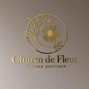 Może przedstawiać: Złote logo dla "Charm de Fleur Flower Boutique" na neutralnym tle. Logo przedstawia okrągły wz&oacute;r z kwiatem i liśćmi. Tekst jest w kroju pisma szeryfowego.