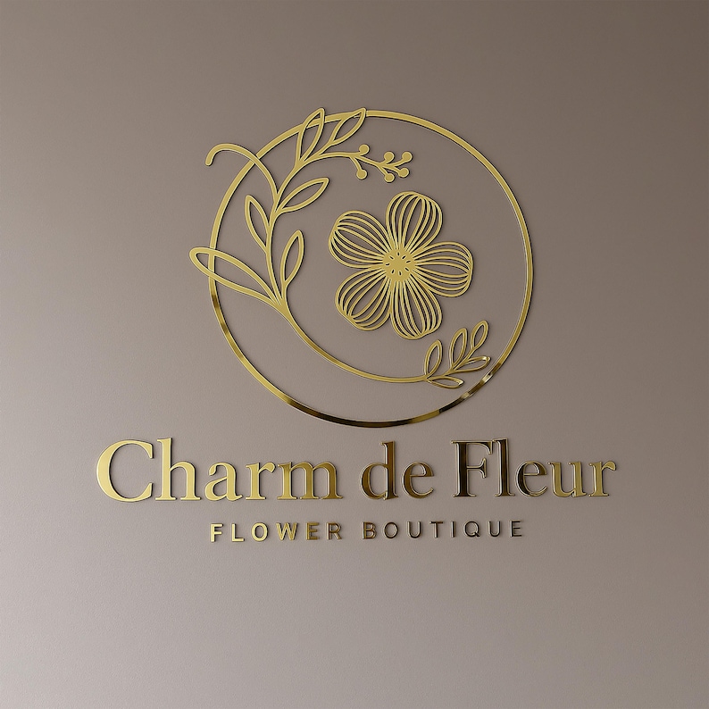 Może przedstawiać: Złote logo dla "Charm de Fleur Flower Boutique" na neutralnym tle. Logo przedstawia okrągły wz&oacute;r z kwiatem i liśćmi. Tekst jest w kroju pisma szeryfowego.