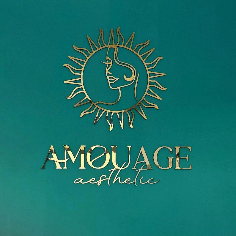 Może przedstawiać: Złote logo na turkusowym tle. Logo przedstawia słońce z profilem kobiety pośrodku. Poniżej znajduje się napis "AMOUAGE" i słowo "aesthetic" w ozdobnej czcionce.