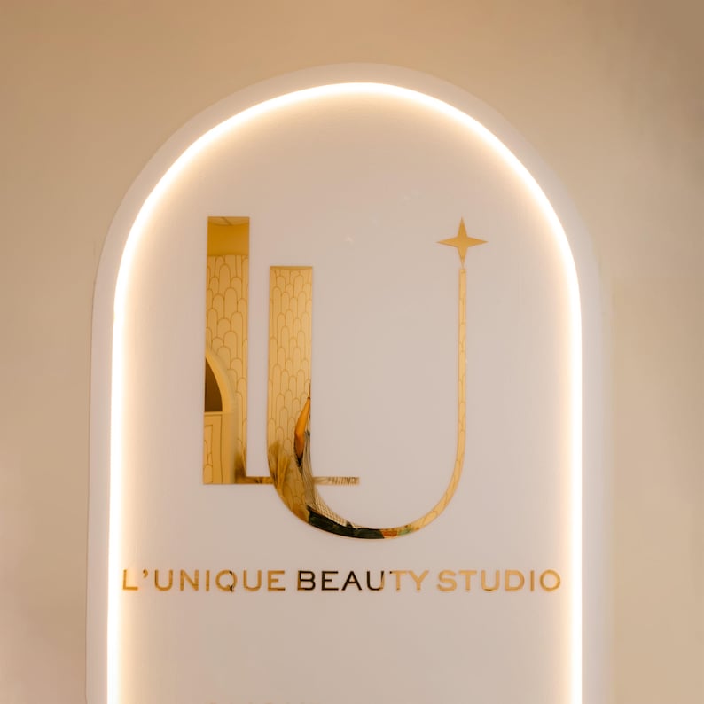 Może przedstawiać: Biały, łukowaty szyld ze złotymi akcentami i napisem "L'UNIQUE BEAUTY STUDIO". Projekt zawiera stylizowane złote elementy architektoniczne i gwiazdę. Szyld jest oświetlony miękkim, ciepłym światłem.