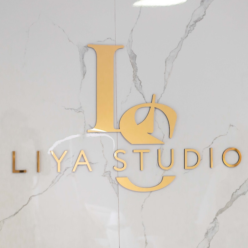 Może przedstawiać: Złote logo "LIYA STUDIO" na białej marmurowej ścianie. Logo zawiera stylizowane litery, z literami "L" i "I" w większym rozmiarze czcionki. Litery są metaliczne i odbijają światło, tworząc nowoczesną i elegancką estetykę.