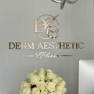May include: A gold-toned, reflective wall sign featuring the letters DA inside a circular frame, with the text DERM AESTHETIC below and the word Clinic in script font, flanked by two horizontal lines, mounted on a plain light grey wall.