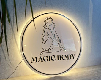 May include: A circular LED wall sign featuring a black line art illustration of a seated figure and the text MAGIC BODY in black capital letters. The sign has a white translucent face with a glowing warm light border and a thin black outer frame.