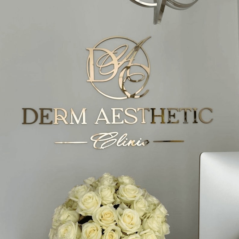 Może przedstawiać: Złoty szyld na ścianie z napisem "DERM AESTHETIC Clinic". Znak zawiera okrągłe logo i linię pod nazwą kliniki. Poniżej znajduje się bukiet białych r&oacute;ż.