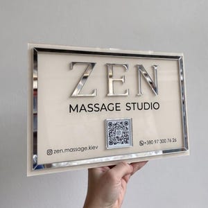 Op de afbeelding: Een rechthoekig bord met een crèmekleurige achtergrond en een zilveren spiegelrand. Het bord bevat de verhoogde zilveren tekst ZEN MASSAGE STUDIO, een centrale QR-code, het Instagram-account zen.massage.kiev en het telefoonnummer +380 97 300 76 26.