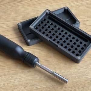 Puede incluir: Un destornillador negro con una punta plateada y una caja de almacenamiento negra con un interior de espuma que contiene múltiples compartimentos.