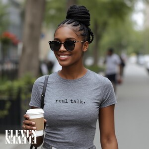 May include: A woman wearing a gray t-shirt with the text "real talk." She is wearing sunglasses and holding a coffee cup. Her hair is styled in a bun. The logo "ELEVEN ELEVEN" is visible at the bottom left.