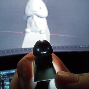May include: A black and green keycap with a glowing face design is held in front of a 3D rendering of a white sculpture. The keycap has a cute, cartoonish design. The background is a computer screen.
