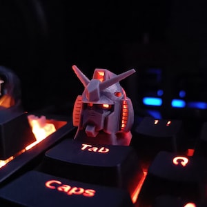 May include: A red and black keyboard with a 3D printed robot head on the Tab key. The keyboard has red backlighting and the robot head is lit up with red lights.