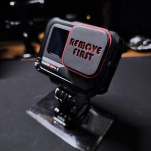 Könnte beinhalten: Eine schwarze Action-Kamera mit roter und schwarzer Schutzabdeckung mit dem roten Schriftzug "REMOVE FIRST". Die Kamera ist auf einem transparenten, rechteckigen Ständer vor dunklem Hintergrund montiert.