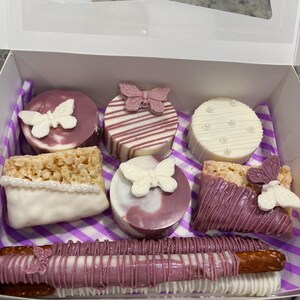 以下が含まれることがあります： お菓子のアソートボックス。ライスクリスピー、チョコレートでコーティングされたプレッツェル、丸いクッキーなど。お菓子は白と紫のアイシングと蝶の形をしたキャンディーで飾られています。箱には紫と白のストライプの裏地が付いています。