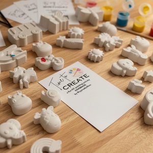 May include: A collection of unpainted plaster figurines in various shapes, including animals, vehicles, and a rainbow. Paint pots in multiple colors, paintbrushes, and cards with text are also present, all arranged on a wooden surface.