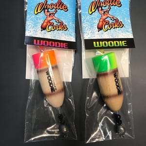 May include: Two fishing corks in clear plastic bags. Each cork has a wooden body with a neon orange or green top. The packaging features the text "Woodie Corks" and a pin-up style graphic on a blue background.
