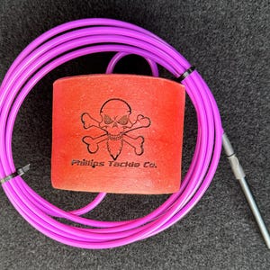 May include: A red and black fishing lure with a skull and crossbones design and the text "Phillips Tackle Co.". The lure is attached to a pink braided fishing line.