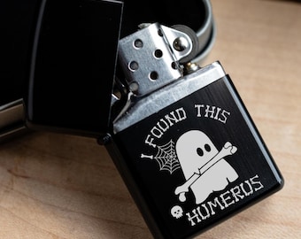 Könnte beinhalten: Schwarzes Feuerzeug mit dem Text "I FOUND THIS HOMERUS" und einer Geistergrafik. Das Feuerzeug ist geöffnet und zeigt das Metallinnere. Das Feuerzeug liegt auf einer Holzoberfläche.