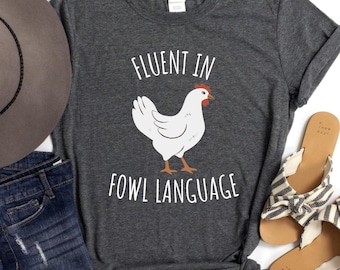 May include: Dark gray t-shirt with the text "FLUENT IN FOWL LANGUAGE" and a white chicken graphic. The shirt is paired with denim shorts, a gray hat, and sandals with black and white striped bows.