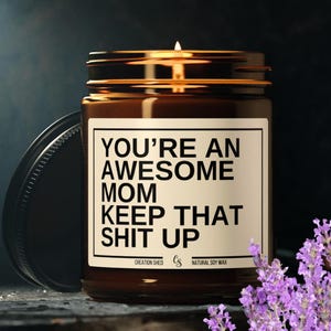 May include: A brown glass candle jar with a black label that reads "You're an Awesome Mom Keep That Shit Up". The candle is surrounded by lavender flowers.