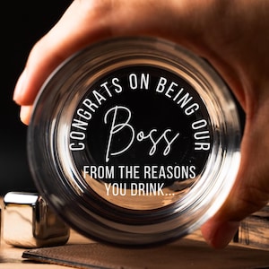 May include: A clear glass with the text "Congrats on being our Boss from the reasons you drink..." etched on the bottom. The glass is being held by a hand.