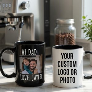 May include: Two ceramic coffee mugs. One black mug features the text "#1 DAD" and a photo of a man and child, with the text "LOVE, JAYLEE". The other mug is white with black trim and the text "YOUR CUSTOM LOGO OR PHOTO".