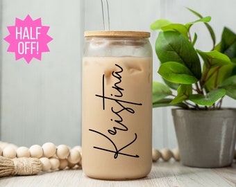 May include: A personalized glass with a bamboo lid and a clear straw. The glass is filled with a light brown beverage and has the name "Kristina" written on it in black.