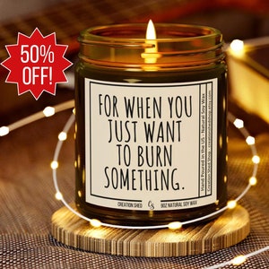 May include: A brown glass candle with a white label that reads "For when you just want to burn something." The candle is lit and sitting on a wooden coaster.