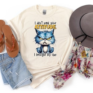 Op de afbeelding: Een beige T-shirt met een blauwe kat cartoon afbeelding en de tekst "I don't need your attitude I brought my own". Het shirt ligt op een witte ondergrond met een bruine hoed, een paar bruine sandalen en een bloemen sjaal.