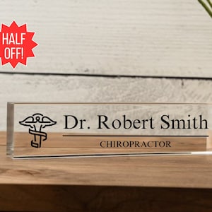 May include: Clear acrylic desk nameplate with a black medical symbol, the text "Dr. Robert Smith" and the title "CHIROPRACTOR".