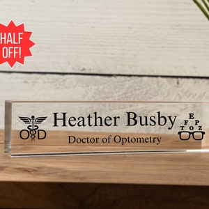 May include: A clear acrylic desk nameplate with black text that reads "Heather Busby, Doctor of Optometry". The nameplate also features a medical symbol with a snake and a staff, and an eye chart with glasses.