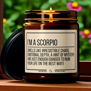 Peut inclure: Une bougie en pot de verre brun allum&eacute;e avec un couvercle noir. La bougie porte une &eacute;tiquette indiquant "I'M A SCORPIO" et d&eacute;crit le parfum. La bougie est sur une surface en bois avec un arri&egrave;re-plan floral vert et rose flou.