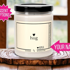 May include: A white soy candle in a clear glass jar with a black lid. The label says "hug" with a heart and "KAYLA" with a heart. The candle is on a wooden surface.