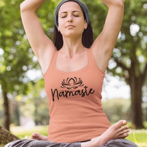 May include: A woman wearing a peach tank top with a black lotus flower and the word "Namaste" printed on the front. She is sitting in a yoga pose with her eyes closed.