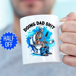 May include: White ceramic mug with the text "DOING DAD SHIT" and a humorous illustration of a skeleton on the toilet, using a phone. The design includes blue watercolor accents and a roll of toilet paper.