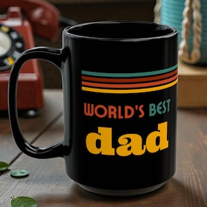 May include: A black ceramic mug with a large handle. The mug features the text "WORLD'S BEST dad" in yellow and a retro-style stripe design in orange, teal, and brown.