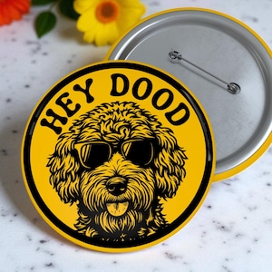 May include: A yellow button with the words "HEY DOOD" in black, featuring a black and yellow illustration of a dog wearing sunglasses. The button has a silver pin back.