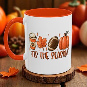 May include: White ceramic mug with an orange interior and handle. The mug features a fall-themed design with a pumpkin spice latte, maple leaf, football, and pumpkin. The text "'Tis the Season" is printed below the graphics.