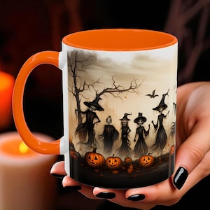 May include: White ceramic mug with an orange interior and handle. The mug features a Halloween-themed illustration of witches, pumpkins, and a spooky tree. The design is set against a muted, atmospheric background.