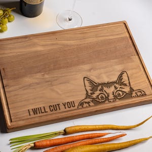 May include: A rectangular wooden cutting board with a juice groove, featuring an engraved illustration of a cat peeking over the edge and the text "I WILL CUT YOU" laser-etched into the surface of the light brown wood grain.