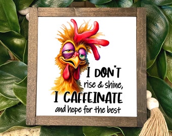 May include: A white sign with a cartoon chicken with a red comb and wattle. The chicken has a tired expression and is looking to the right. The sign says "I don't rise & shine, I caffeinate and hope for the best".