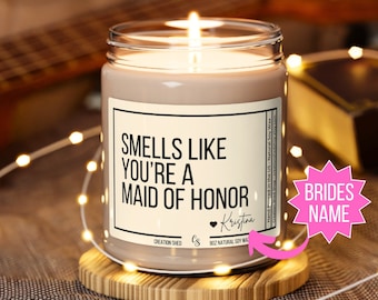 Könnte beinhalten: Eine braune Sojawachskerze mit einem weißen Etikett, auf dem "Smells Like You're a Maid of Honor" mit einem Herz und dem Namen Kristina steht. Die Kerze steht auf einem Holzuntersetzer und ist von weißen Lichterketten umgeben.