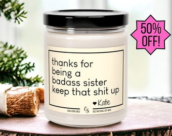 May include: A white candle with a black lid and a label that reads "thanks for being a badass sister keep that shit up Katie". The candle is on a wooden surface.