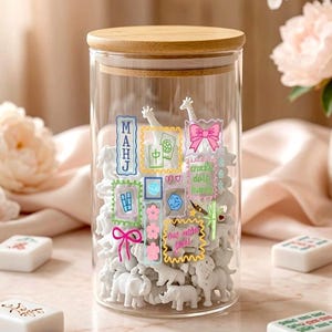 May include: A clear glass cylindrical storage jar with a light wood lid, filled with small white animal figurines. The exterior of the glass features colourful, whimsical sticker-style illustrations of bows, flowers, and Mahjong-related symbols.