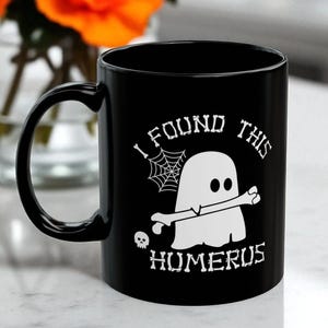 Op de afbeelding: Een zwarte keramische mok met een witte afbeelding van een geest die een bot vasthoudt. De tekst I FOUND THIS HUMERUS staat in een bot-achtig lettertype. Een klein spinnenweb en een schedel maken het griezelige ontwerp op de mok compleet.