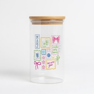 May include: A clear glass storage jar with a light brown wooden lid. The exterior features colourful illustrations of mahjong tiles, a pink bow, a bamboo stalk, and text that reads "MAHJ", "cracks, dots, bams", and "one more game" in a stamp-like design.