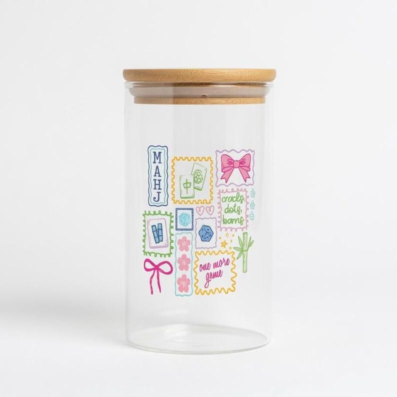 May include: A clear glass storage jar with a light brown wooden lid. The exterior features colourful illustrations of mahjong tiles, a pink bow, a bamboo stalk, and text that reads "MAHJ", "cracks, dots, bams", and "one more game" in a stamp-like design.