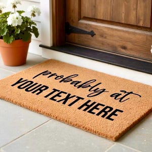 May include: A rectangular coir doormat with the text "probably at YOUR TEXT HERE" in black script. The mat is light brown and sits on a tiled surface in front of a wooden door. Potted plants with white flowers are on either side.
