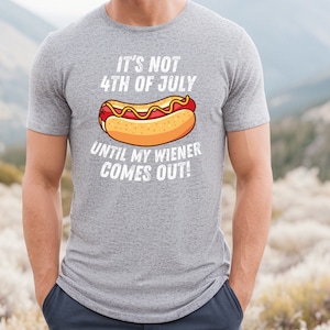 Pode incluir: Uma camiseta cinza com um gr&aacute;fico branco de um cachorro-quente e o texto "It's not 4th of July until my wiener comes out!"