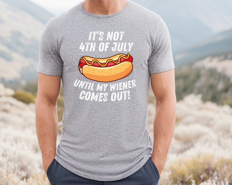 Pode incluir: Uma camiseta cinza com um gr&aacute;fico branco de um cachorro-quente e o texto "It's not 4th of July until my wiener comes out!"