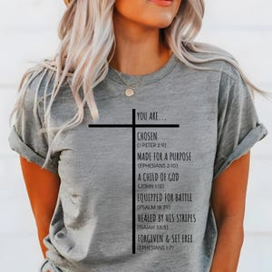 May include: A gray t-shirt with a black cross and the text "You are... Chosen (1 Peter 2:9) Made for a purpose (Ephesians 2:10) A child of God (John 1:12) Equipped for battle (Psalm 18:39) Healed by his stripes (Isaiah 53:5) Forgiven & set free (Ephesians 1:7)"