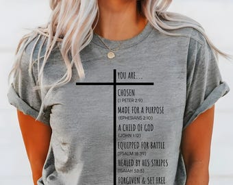 May include: A gray t-shirt with a black cross and the text "You are... Chosen (1 Peter 2:9) Made for a purpose (Ephesians 2:10) A child of God (John 1:12) Equipped for battle (Psalm 18:39) Healed by his stripes (Isaiah 53:5) Forgiven & set free (Ephesians 1:7)"